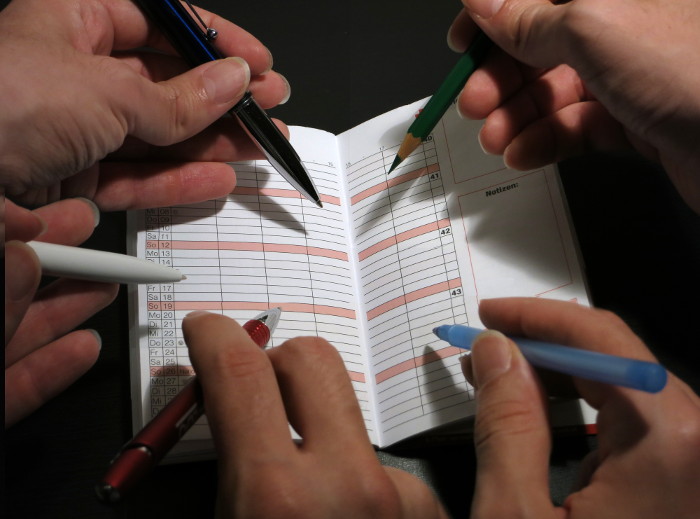fünf Hände mit Stiften wollen auf ein Kalenderblatt schreiben.