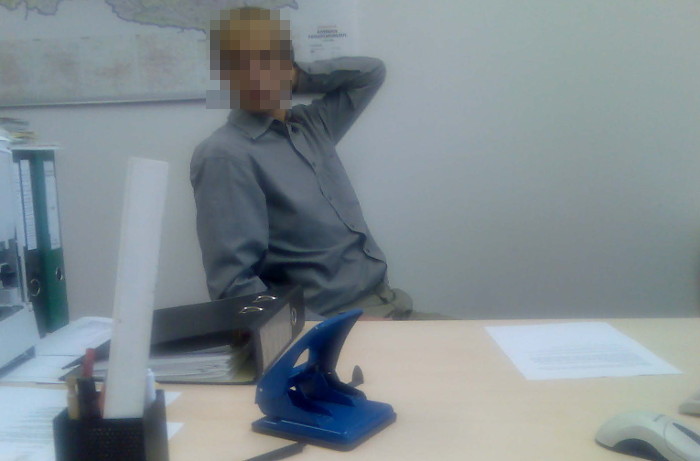 Mann in grauem Hemd hinter Schreibtisch. Das Gesicht ist verpixelt.
